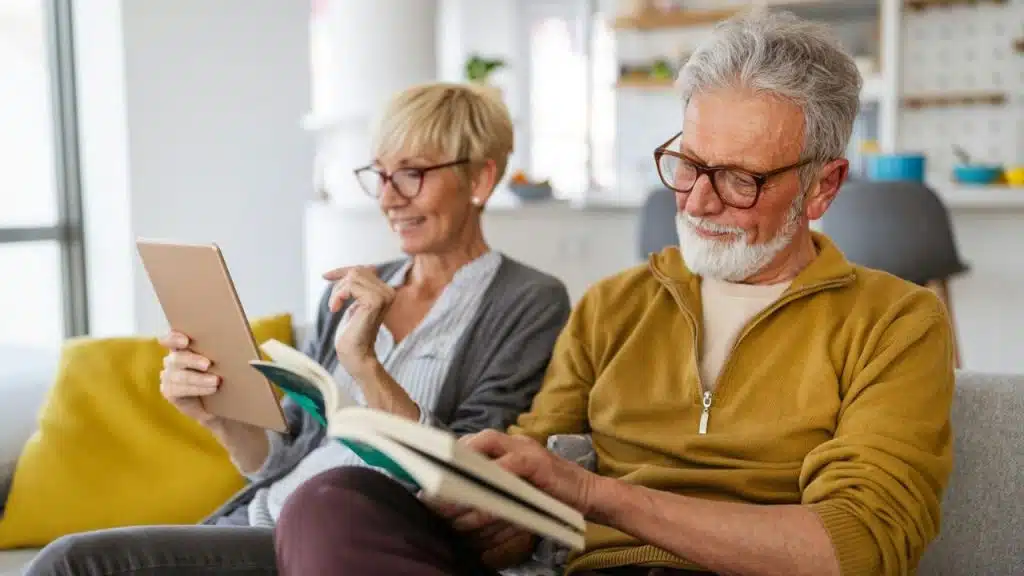 A smiling senior couple reads, with a woman on a tablet and a man with a beard reading a book.