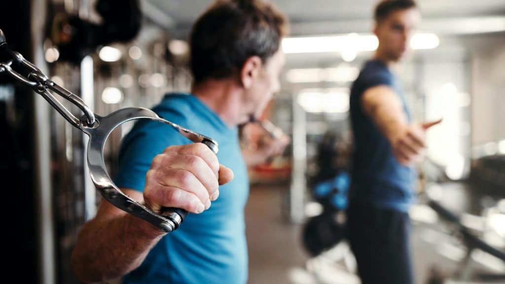 A man using a cable machine at the gym with a trainer nearby.