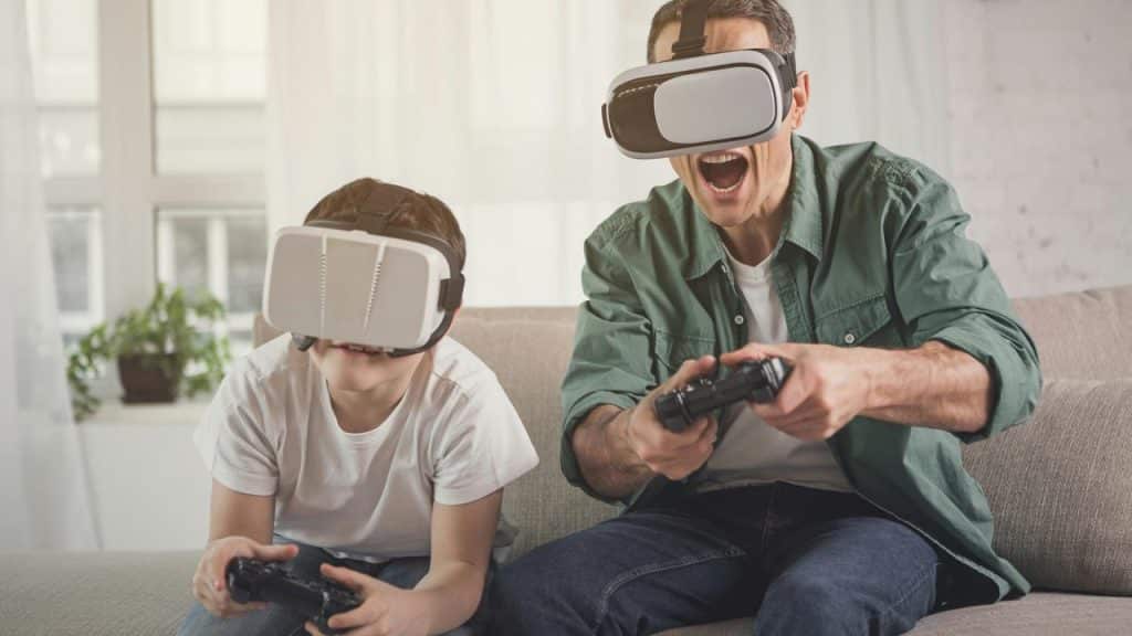 A man and a kid wearing VR headsets and playing video games with controllers on a couch.
