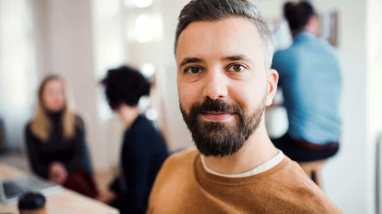 A bearded man in a brown sweater smiles at the camera in an office.