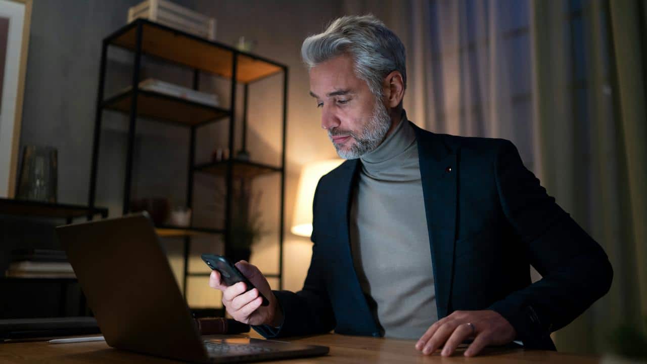 A man in a blazer and turtleneck checking his phone at a desk with a laptop.