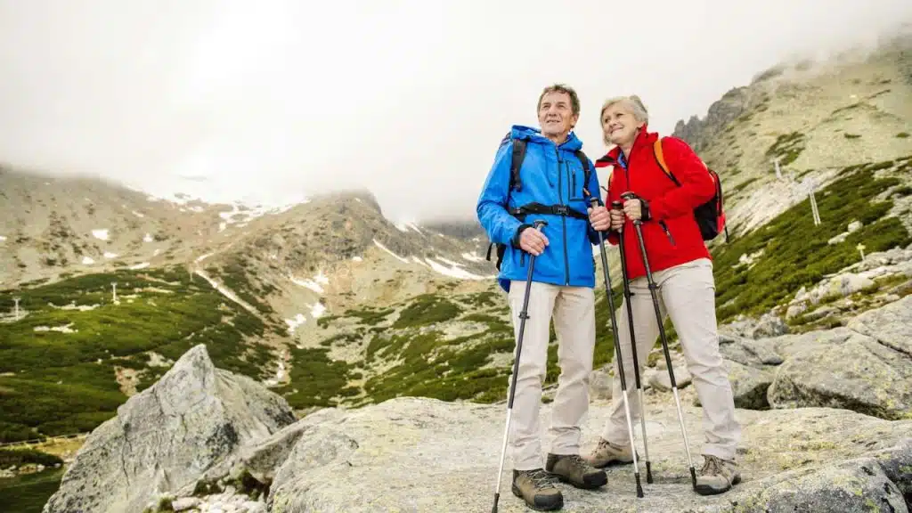 A senior couple, dressed in hiking gear, smiles while standing on a rocky mountain path.