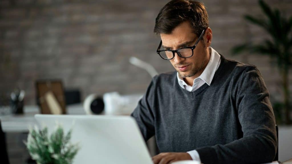A man with dark hair and glasses works intently on a laptop at a desk.