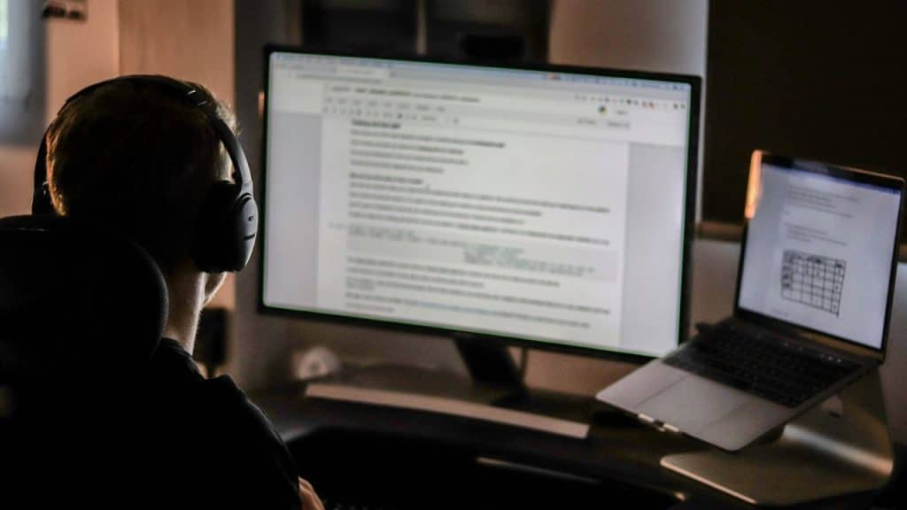 A person with headphones looks at a computer monitor and an open laptop in a dimly lit room.