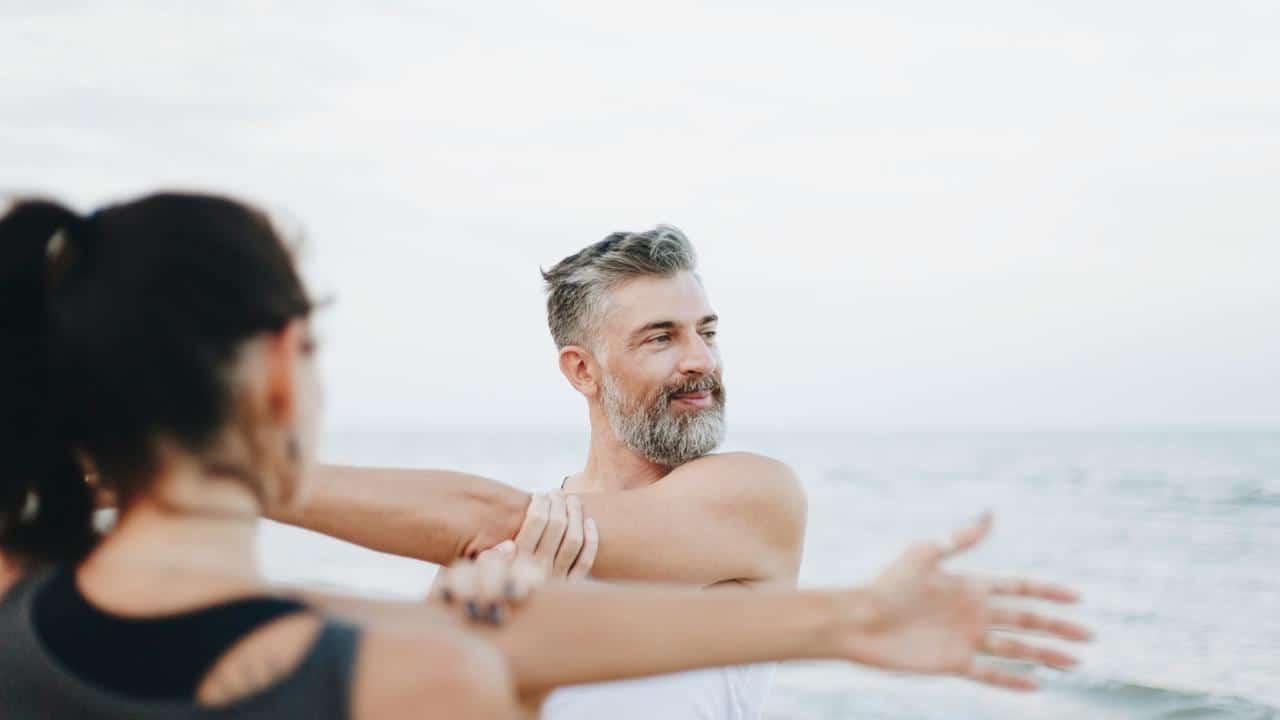 A smiling bearded man stretching his arm at the beach.