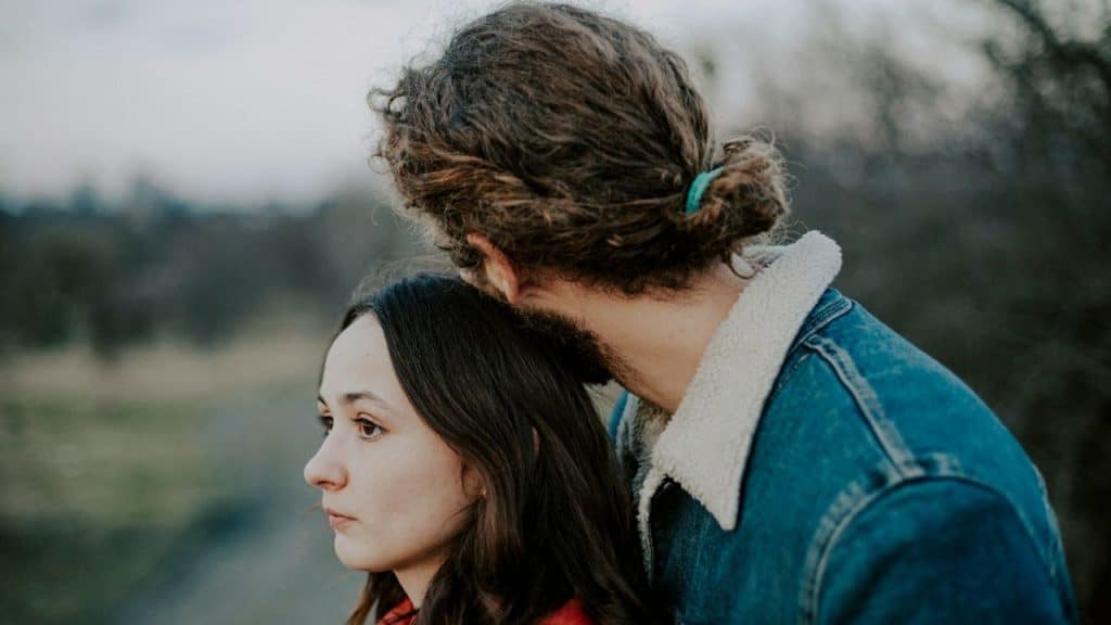 A man with curly hair tied back stands behind a woman with long dark hair.