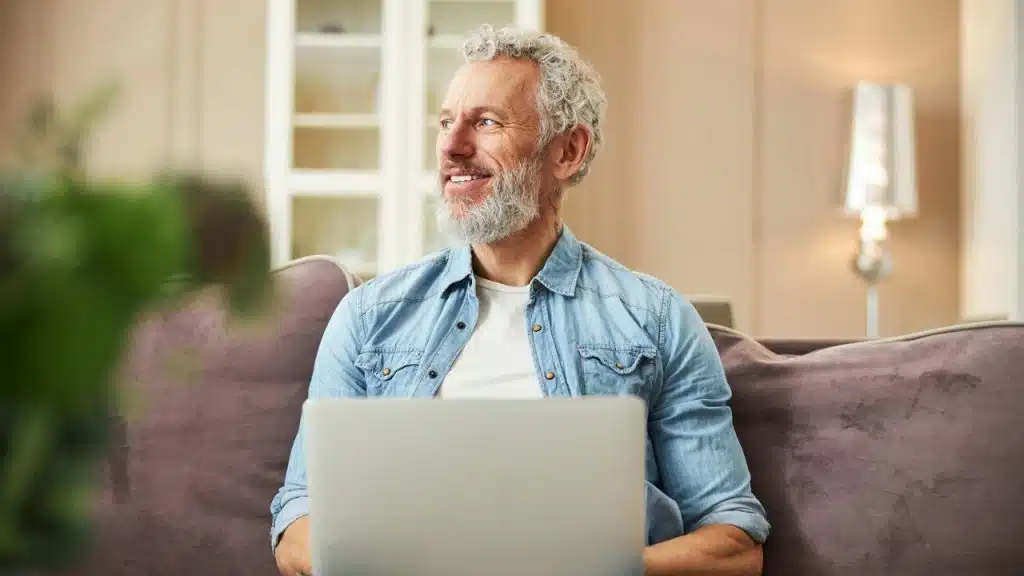 A smiling man with a white beard and glasses sits on a couch, looking to the side with a laptop.