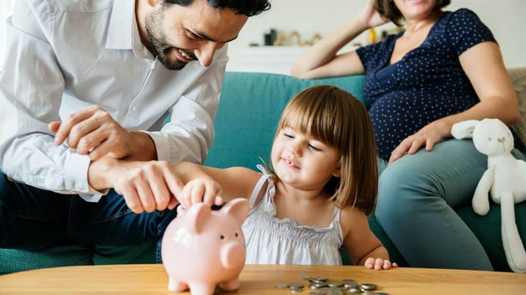 A smiling father helps a young girl put money into a piggy bank, with a woman in the background.