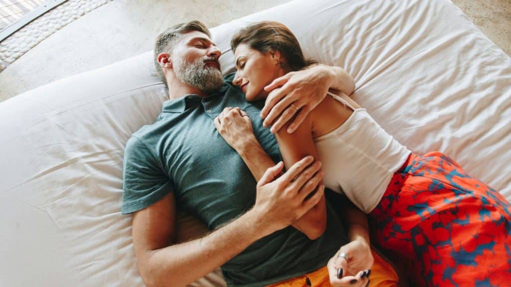 A man and woman embrace in bed, eyes closed, appearing peaceful.
