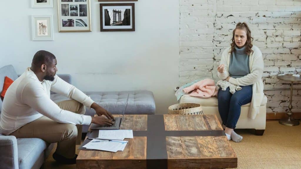 A man works on a laptop while a woman sits on a bench, looking distressed.