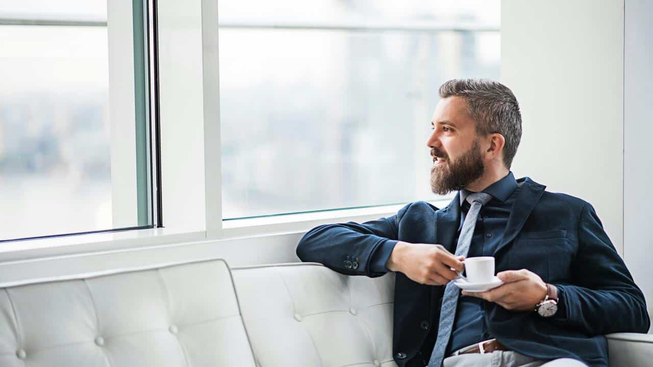 A man in a suit drinks coffee while sitting by a window.