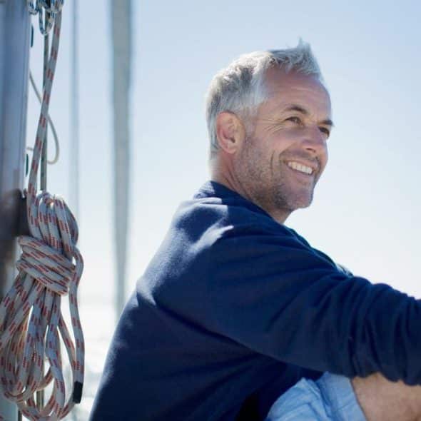 A smiling middle-aged man in a sweater sitting on a sailboat with the ocean behind him.