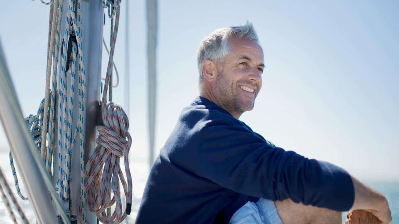 A smiling middle-aged man in a sweater sitting on a sailboat with the ocean behind him.