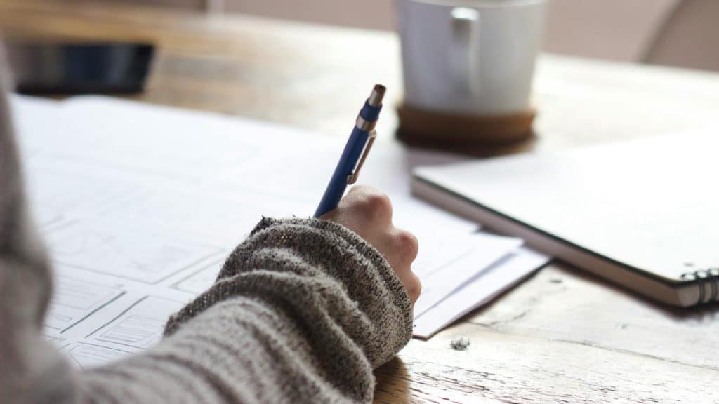 A person writing on paper with a pen at a wooden desk.