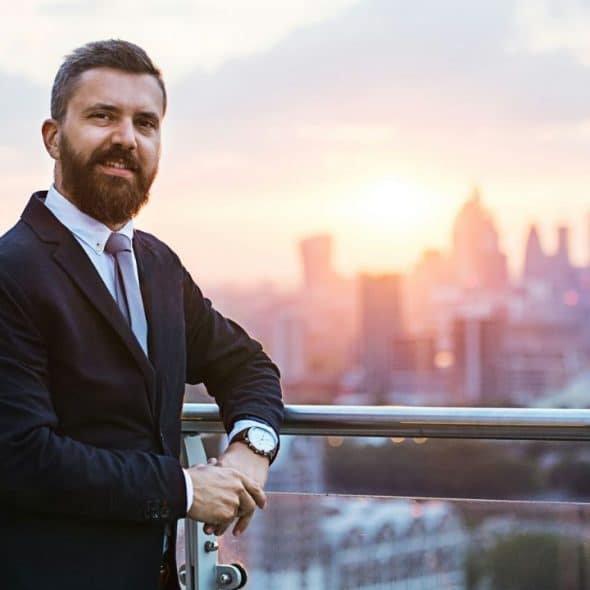 A smiling bearded man in a suit stands on a balcony overlooking a city.