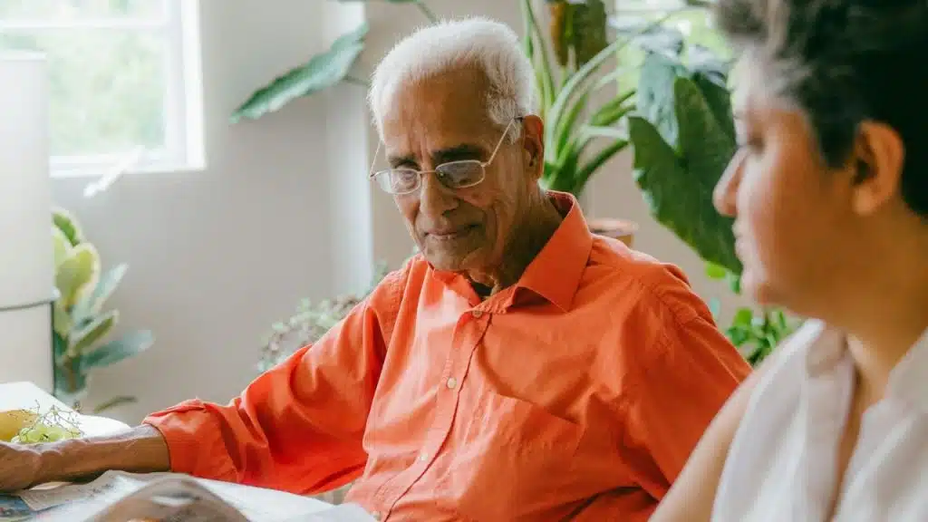 An elderly man in an orange shirt and glasses looks down at a book, with a person beside him.