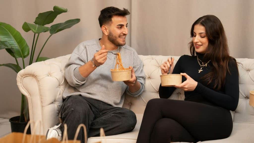 A couple happily shares a meal from takeout containers on a sofa..