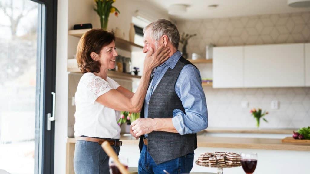 A smiling woman gently touches a man's face in a kitchen setting.