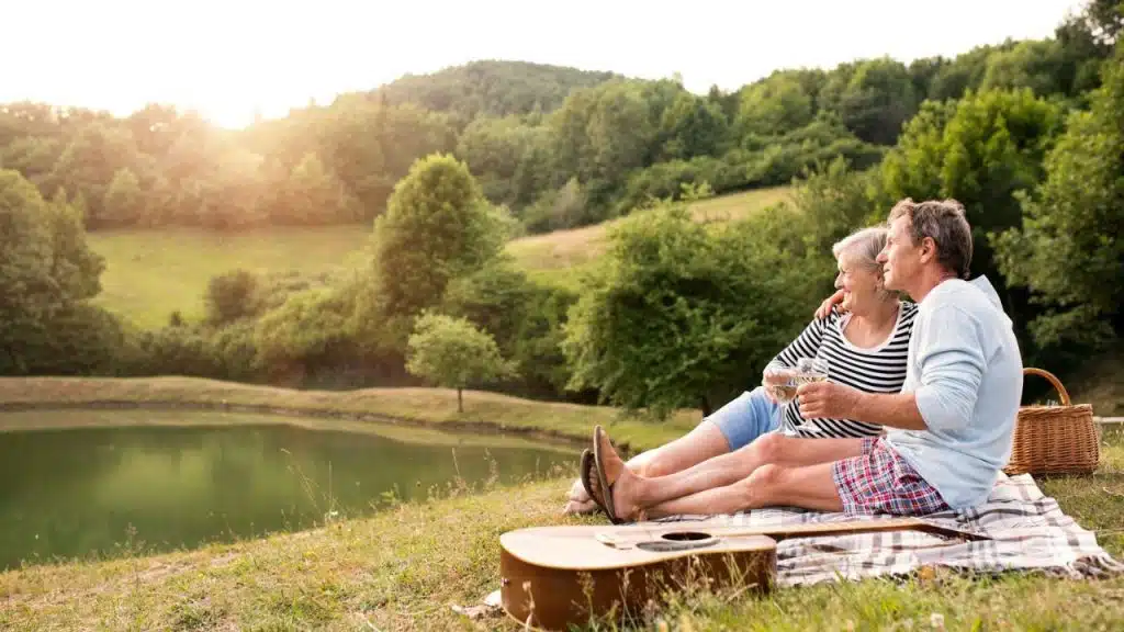 A happy senior couple sits on a blanket by a lake, enjoying a picnic with a guitar nearby.