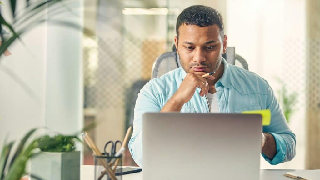 A man with a serious expression, wearing a light blue shirt, looks intently at a laptop screen.