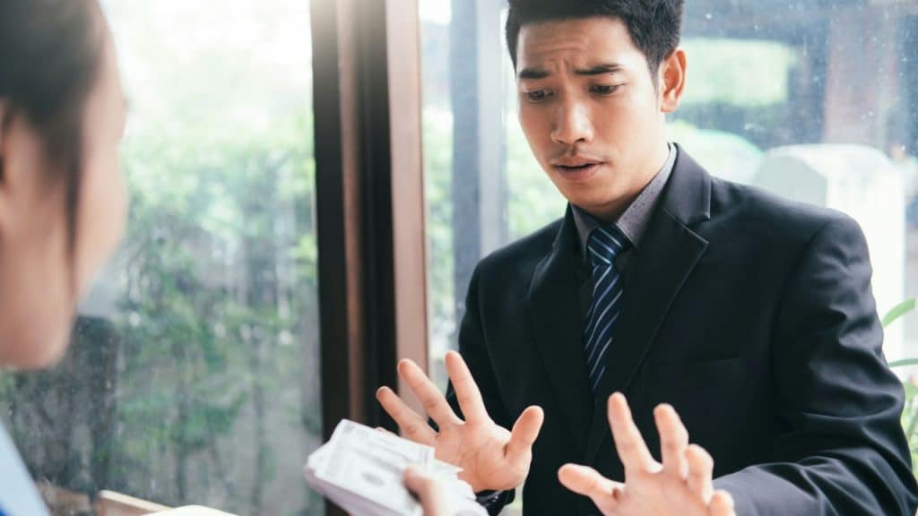 A man in a suit makes a "stop" gesture as someone offers him a stack of money.