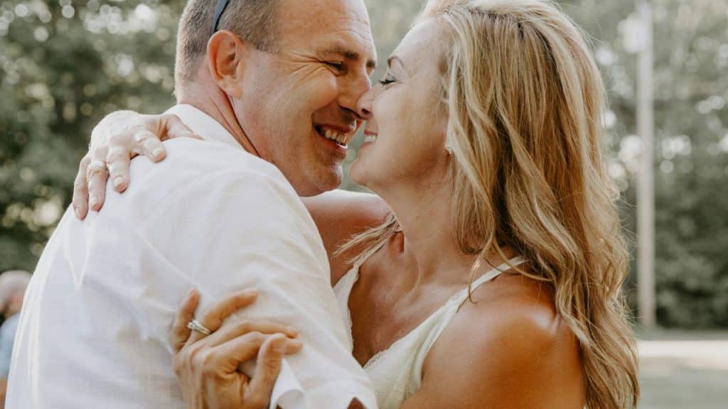 A joyful middle-aged couple in white embracing outdoors, laughing with eyes closed.