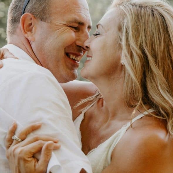 A joyful middle-aged couple in white embracing outdoors, laughing with eyes closed.