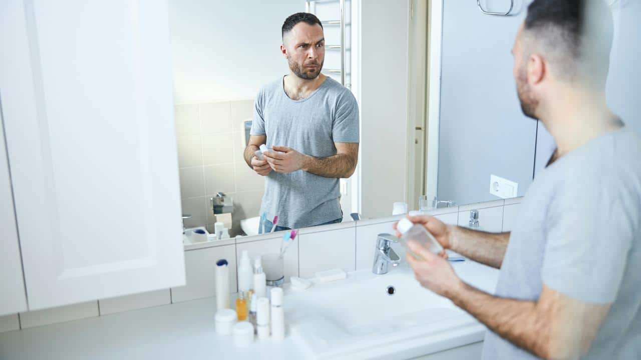 A man in a grey shirt looks at himself in the bathroom mirror.