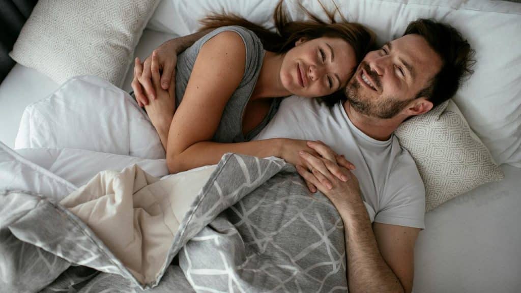 A couple cuddling in bed under a gray blanket.