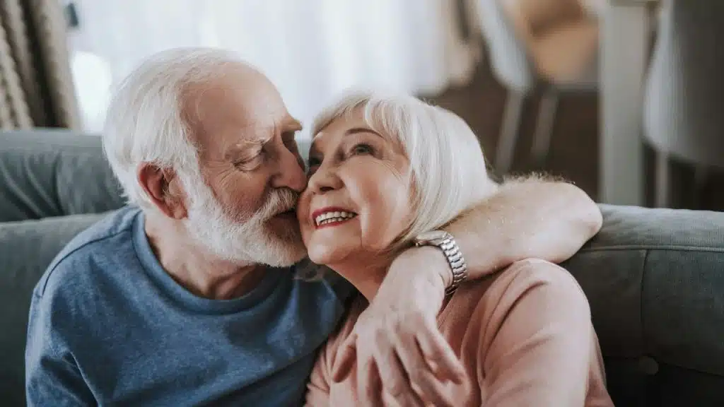 A happy elderly couple embraces on a couch, with the man kissing the woman's cheek.