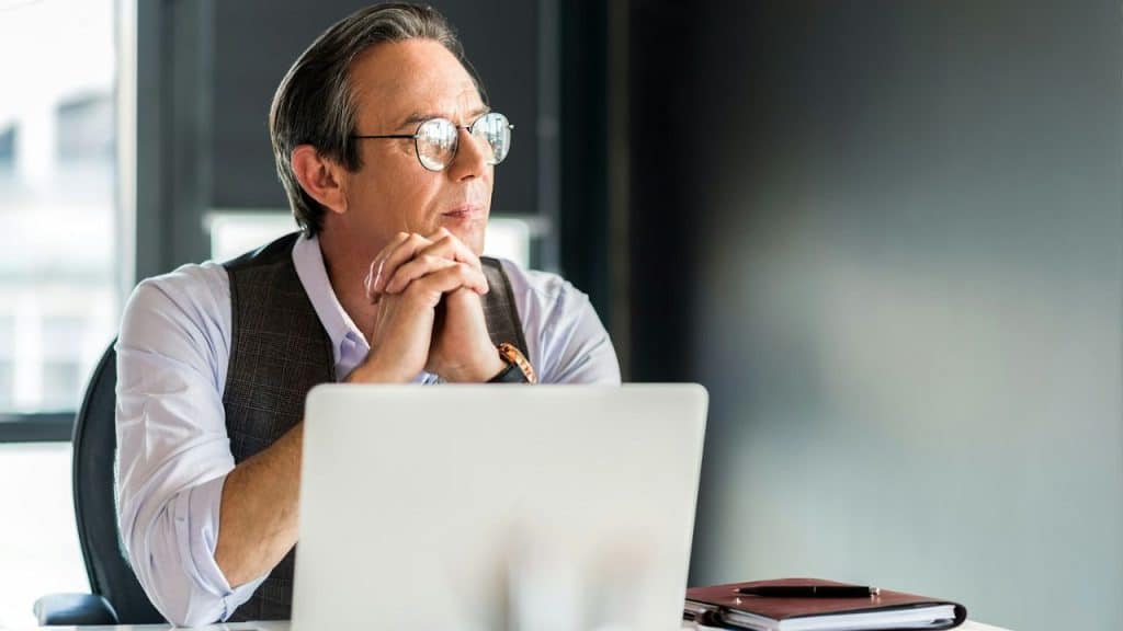 A pensive man with glasses, wearing a white shirt and vest, sits at a desk with a laptop.
