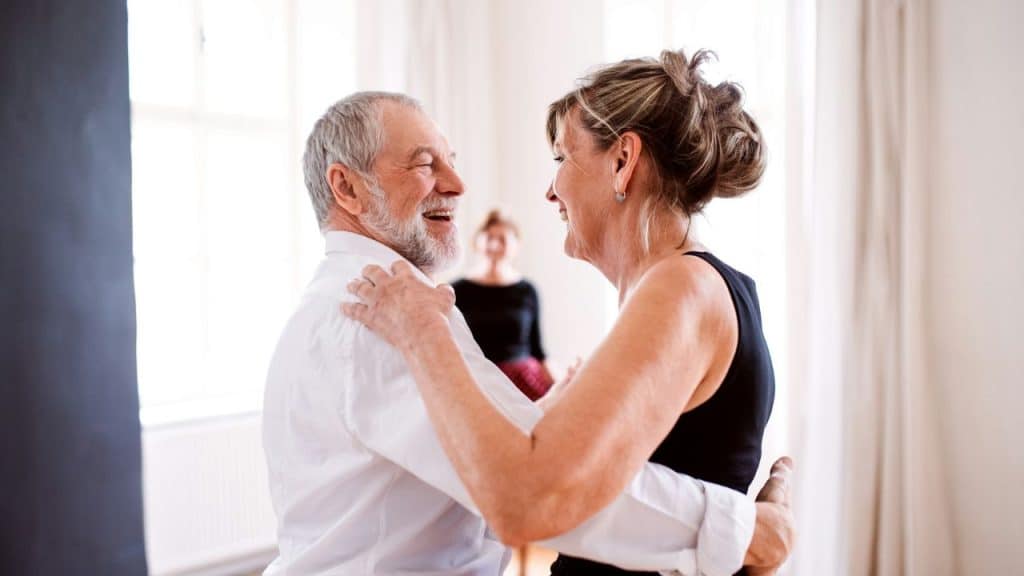 A cheerful older couple in a bright room embraces, seemingly dancing, with a blurry figure in the background.