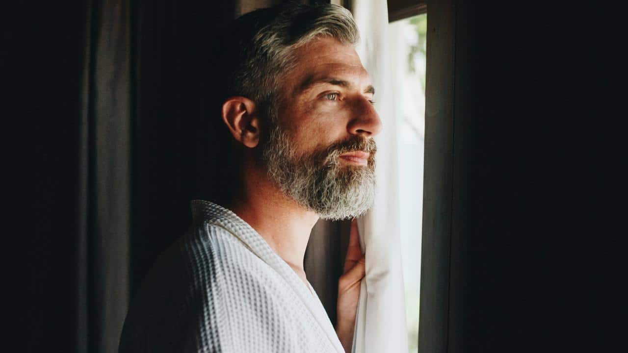 A man in a robe looks out a window with soft light.