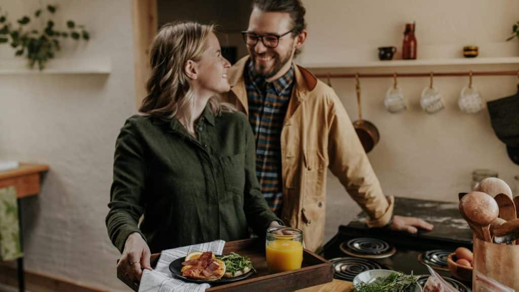 A couple smiling at each other in a kitchen with breakfast on a tray.