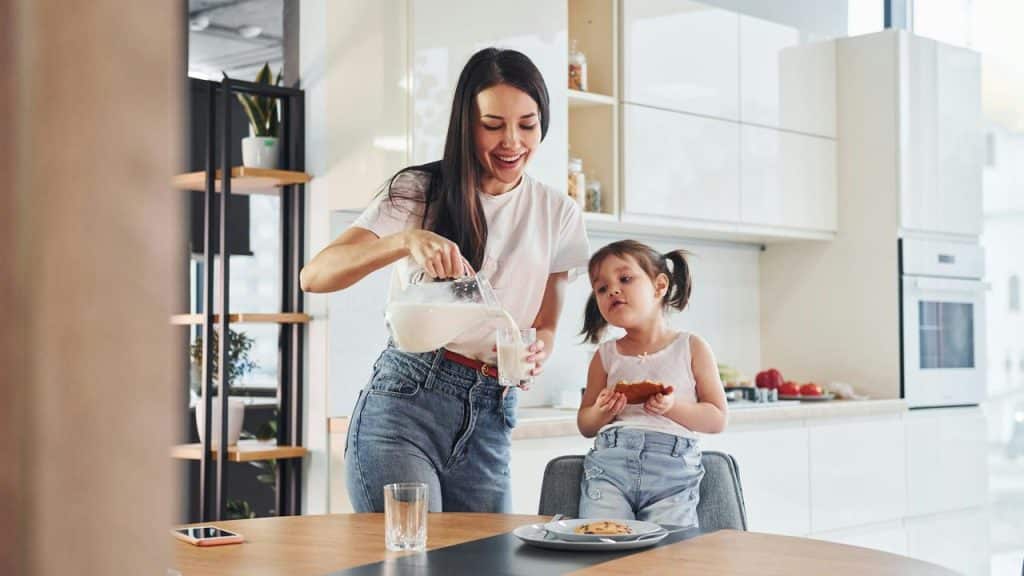 A mother pouring milk while her daughter eats a cookie.