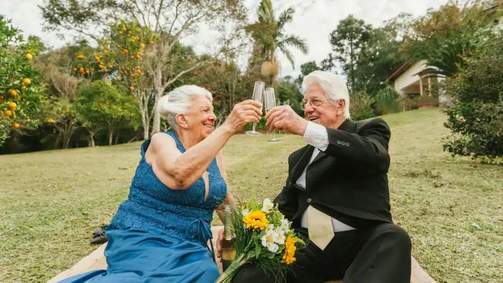 A joyful elderly couple, dressed up, makes a toast with champagne glasses in a grassy outdoor setting.