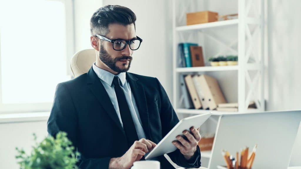 A professional man in a suit and glasses sits at a desk, looking at a tablet with a laptop nearby.