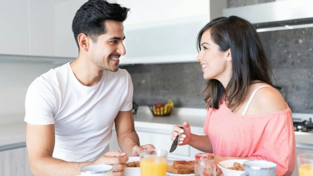 A happy couple shares breakfast at a table, smiling at each other.
