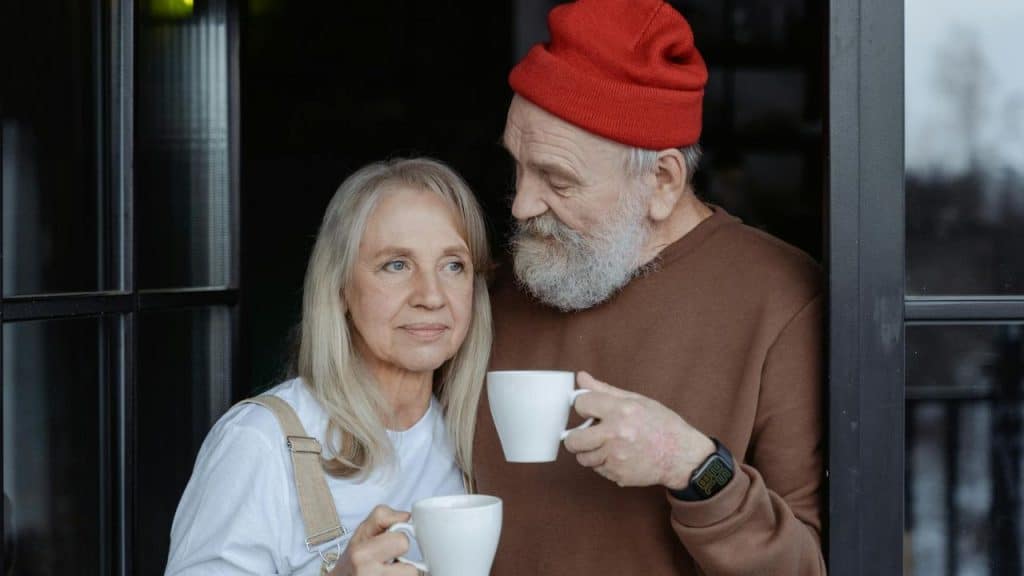A smiling elderly couple holds coffee cups by a window, looking content.