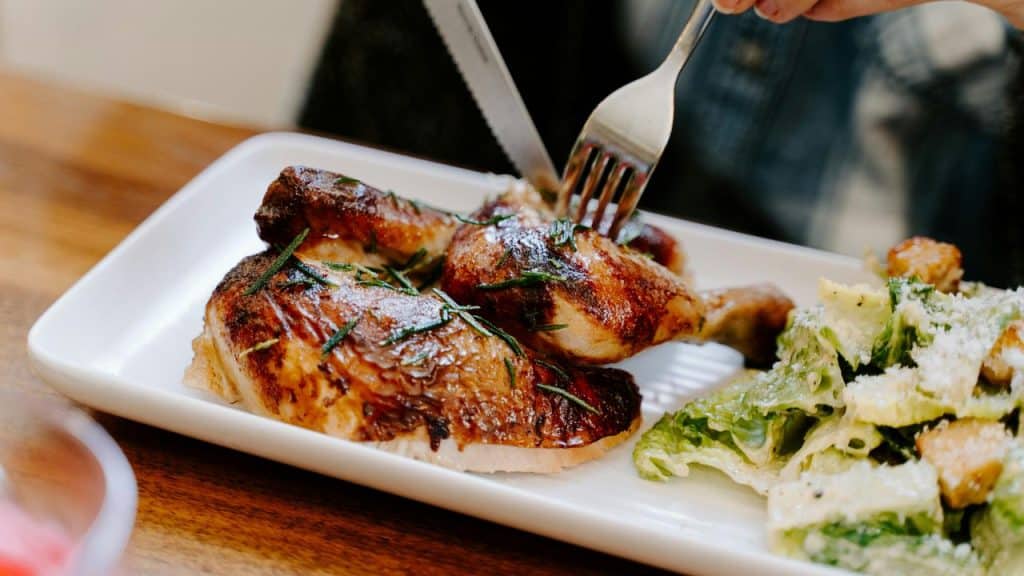 Person using a fork and a knife to cut grilled chicken served in a white rectangular plate