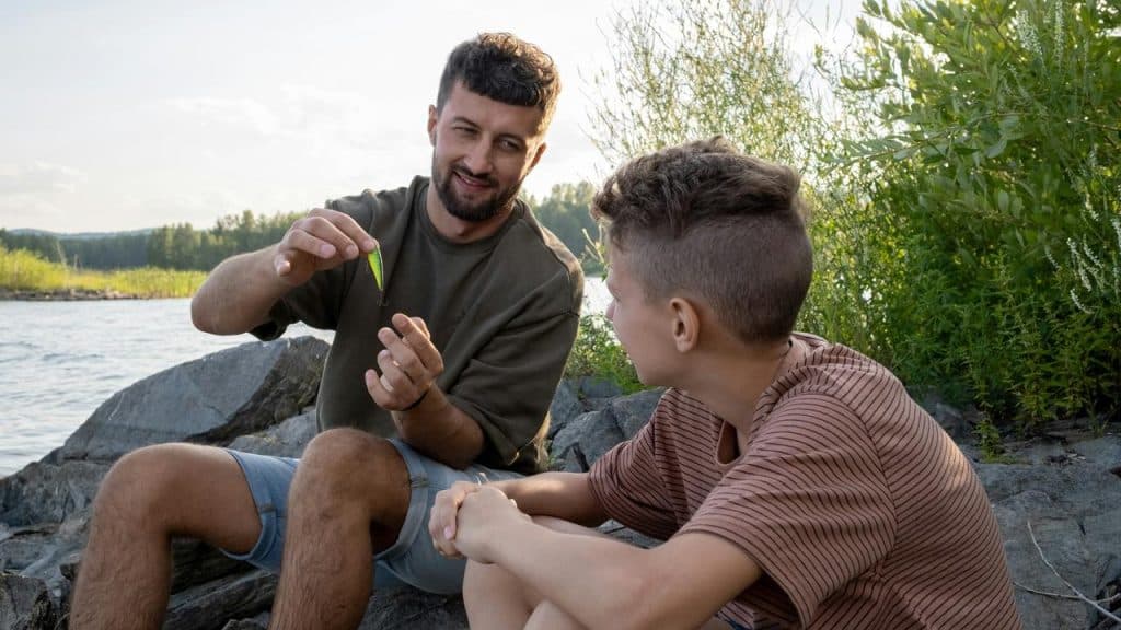 A man showing a fishing lure to a kid by the water.
