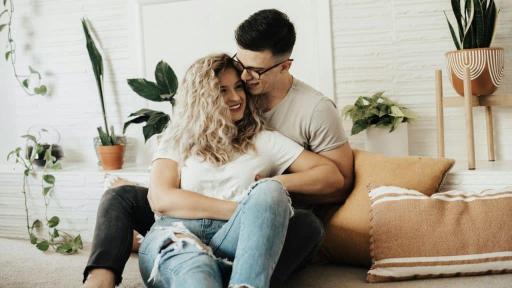 A couple embraces and smiles while sitting on the floor surrounded by plants.