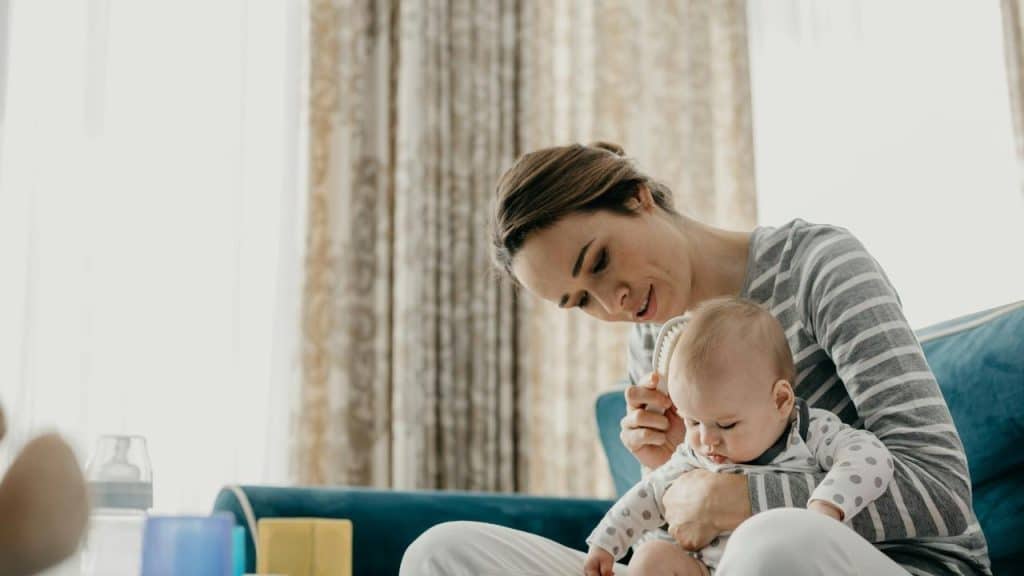 A mother brushing her baby’s hair while sitting on a couch.