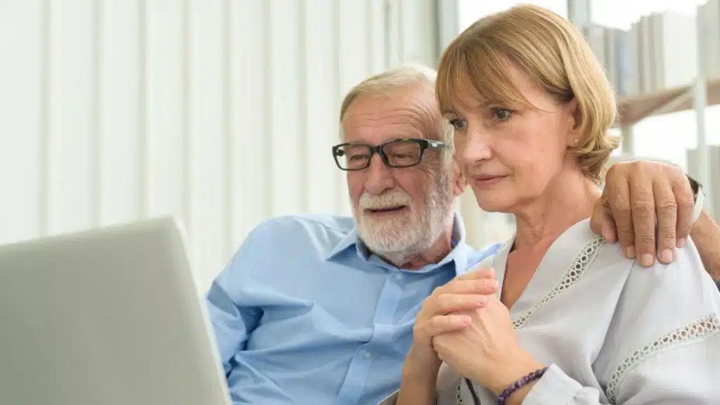 A senior couple sits closely, looking at a laptop, with the man's arm around the woman.