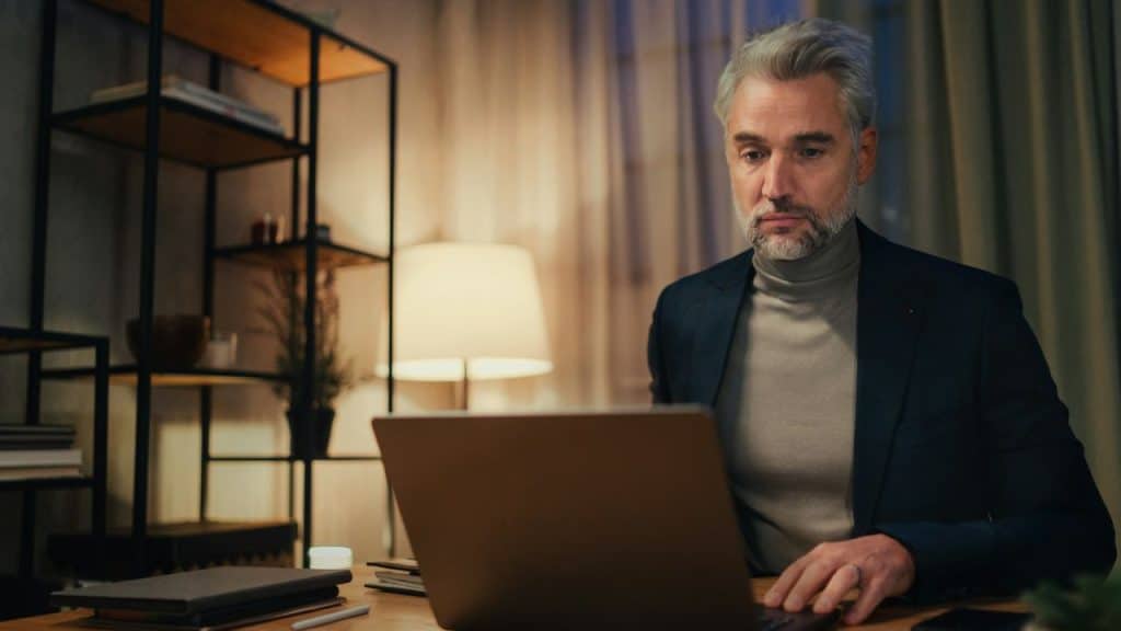 A distinguished man with grey hair and a beard works intently on a laptop at a desk.
