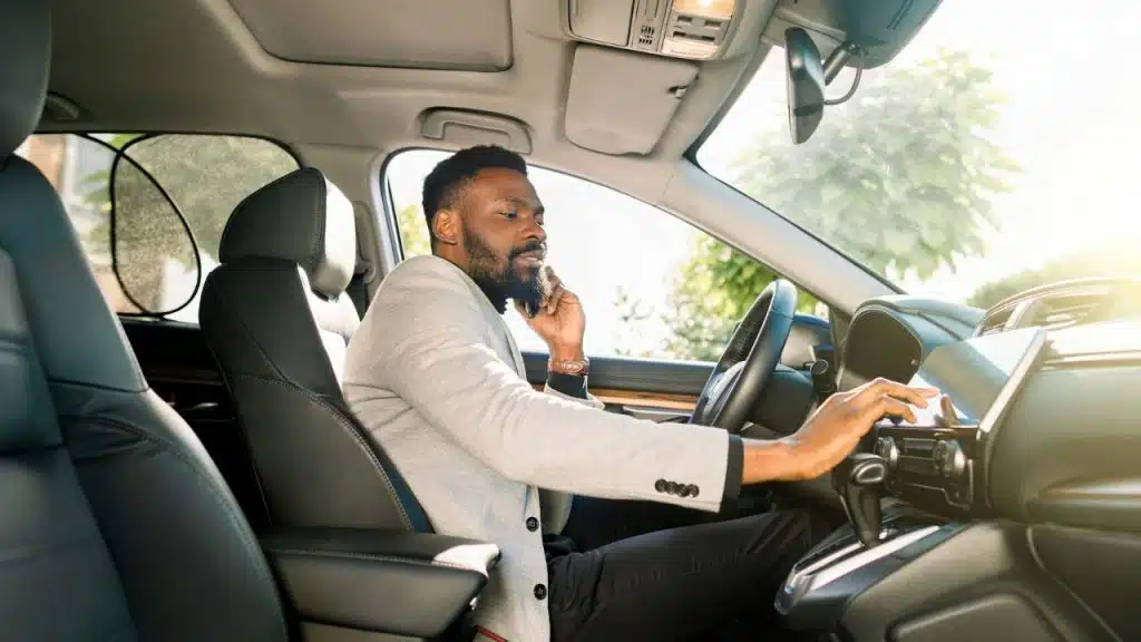 A man talks on the phone while adjusting the car's screen, sunlight streaming in.