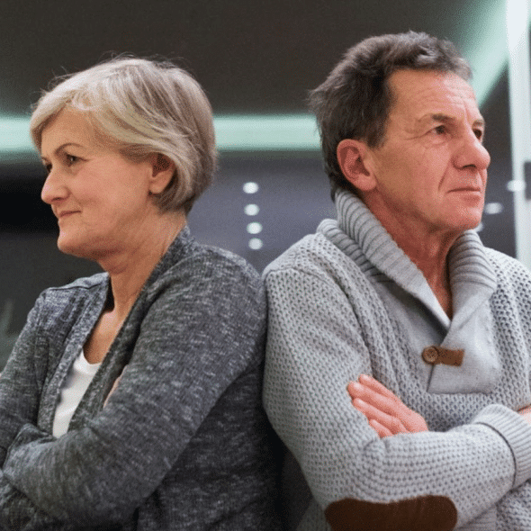 An older man and woman sit back-to-back on a sofa with their arms crossed.