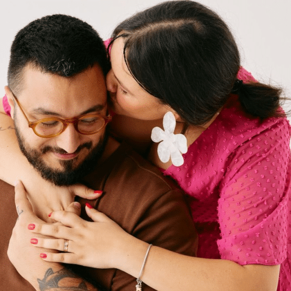 A woman in a magenta dress hugs a man from behind, kissing his cheek.