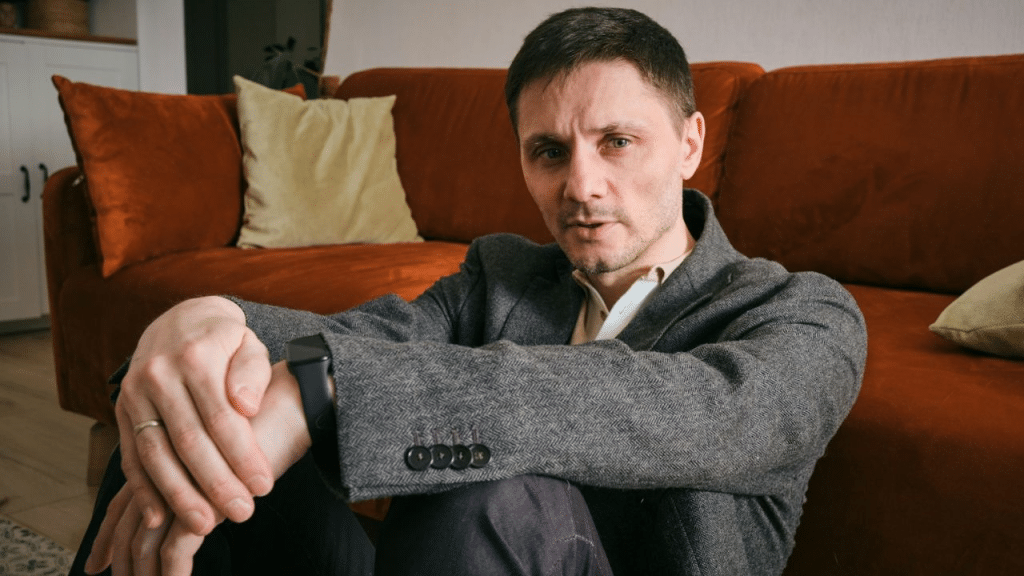 A man in a blazer sits on the floor, leaning against a couch, with a serious expression.
