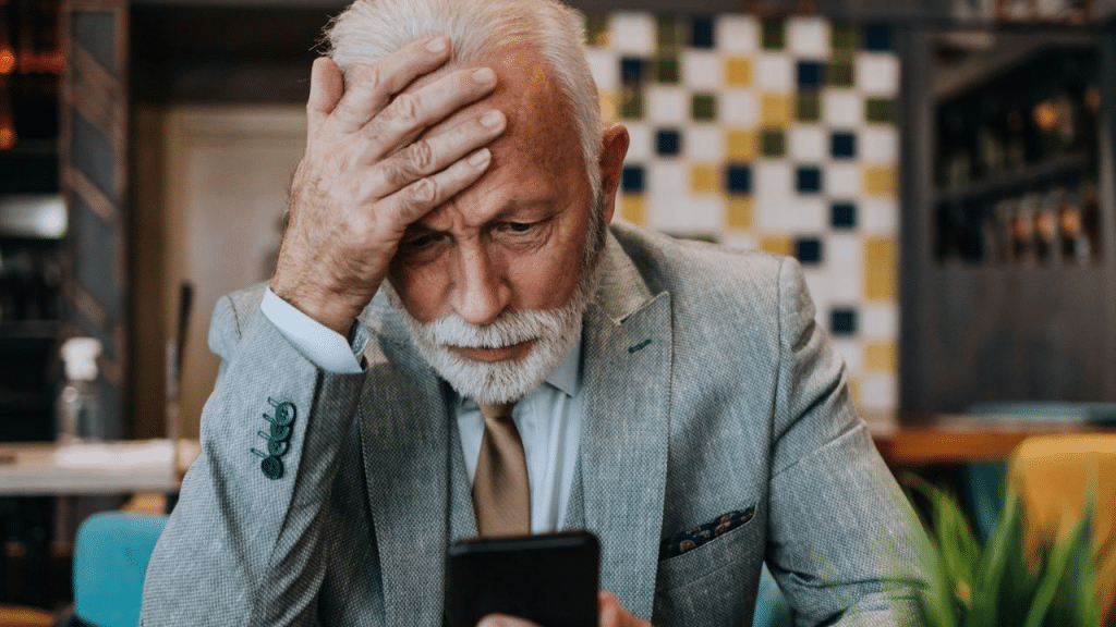 An older man in a blazer looks at his phone with a distressed expression.